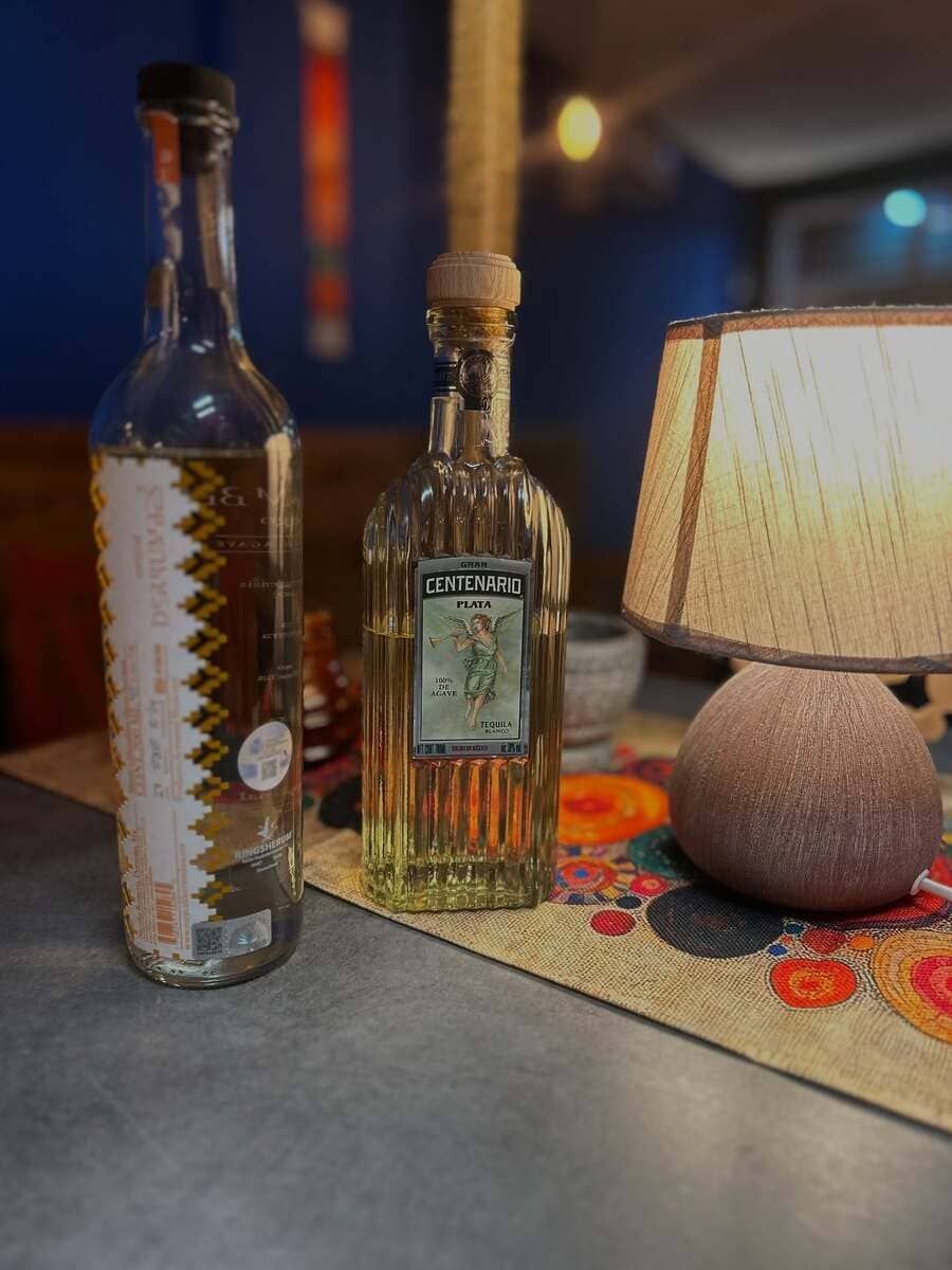 Dos botellas de tequila sobre una mesa, camino de flores, lámpara, tenuemente iluminada.