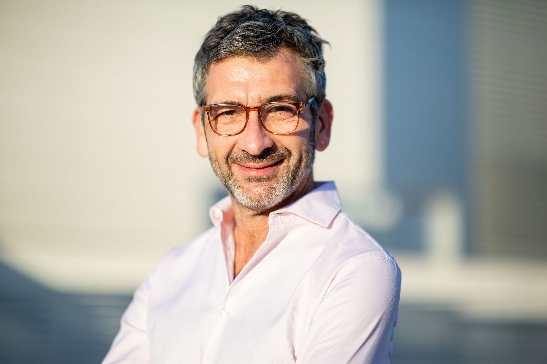 Eine Person mit kurzen, dunklen Haaren und Brille lächelt in einem hellrosa Hemd vor einem verschwommenen Hintergrund.