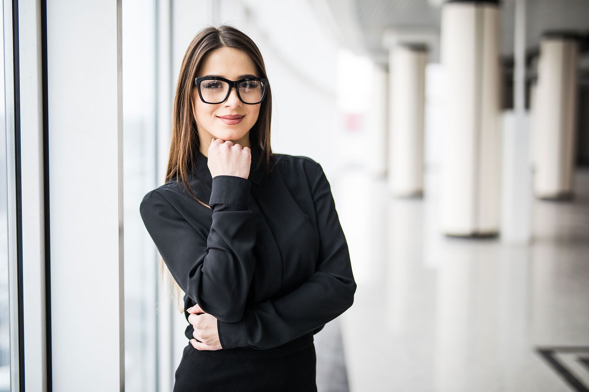 Eine Frau in schwarzem Hemd und Brille posiert in einem hellen Büroflur mit Fenstern.