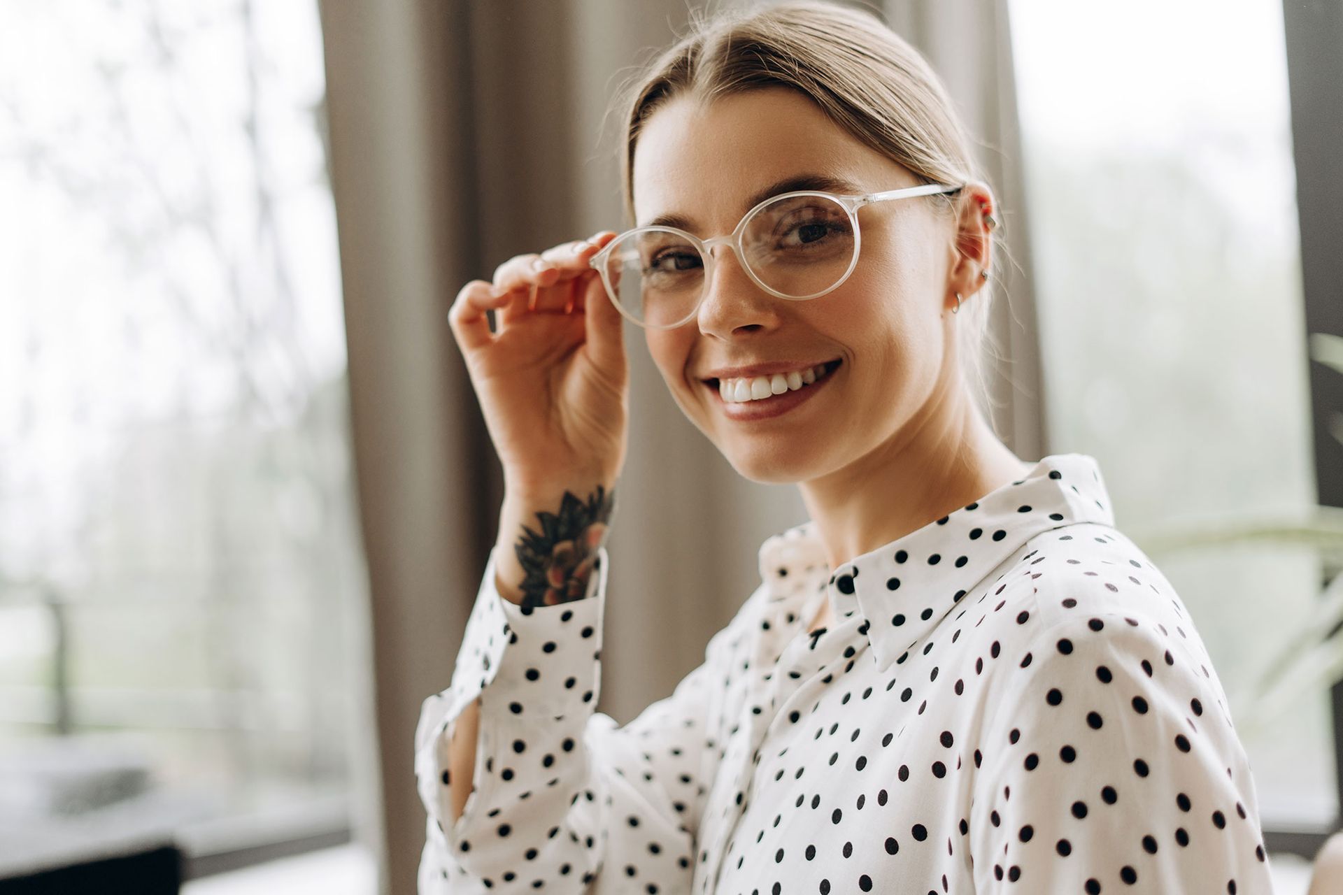 Eine lächelnde Person mit einem Tattoo am Handgelenk trägt ein weißes Hemd und rückt ihre durchsichtige Brille zurecht.