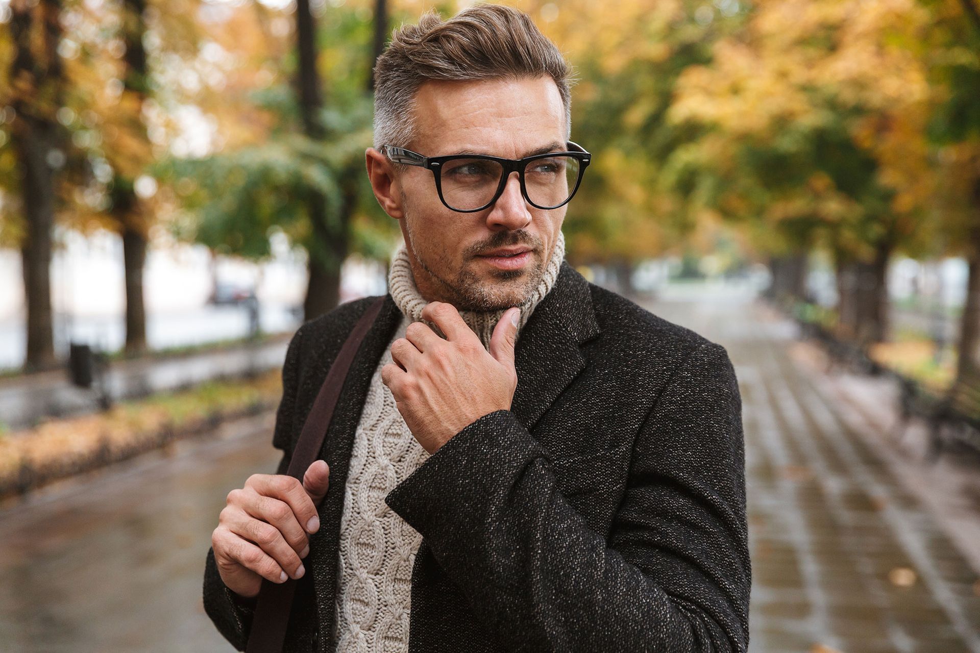 Ein Mann in schwarzem Mantel und Zopfstrickpullover steht im Herbst auf einem von Bäumen gesäumten Gehweg.