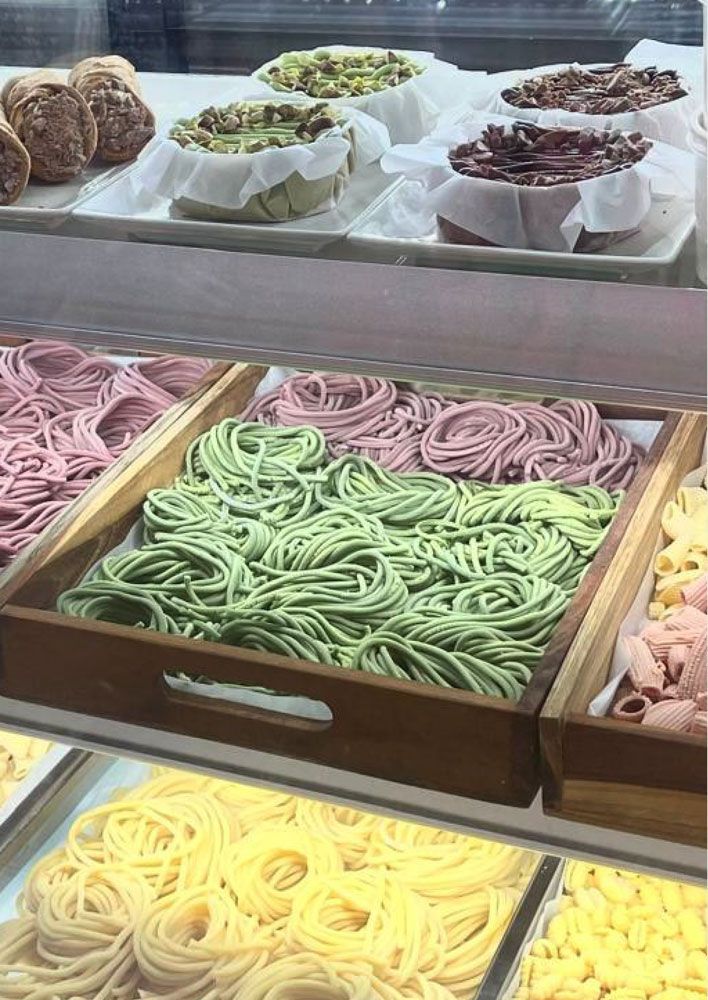 Una vitrina repleta de colorida pasta fresca y pasteles redondos, incluyendo montones de fideos verdes, amarillos y rosas.