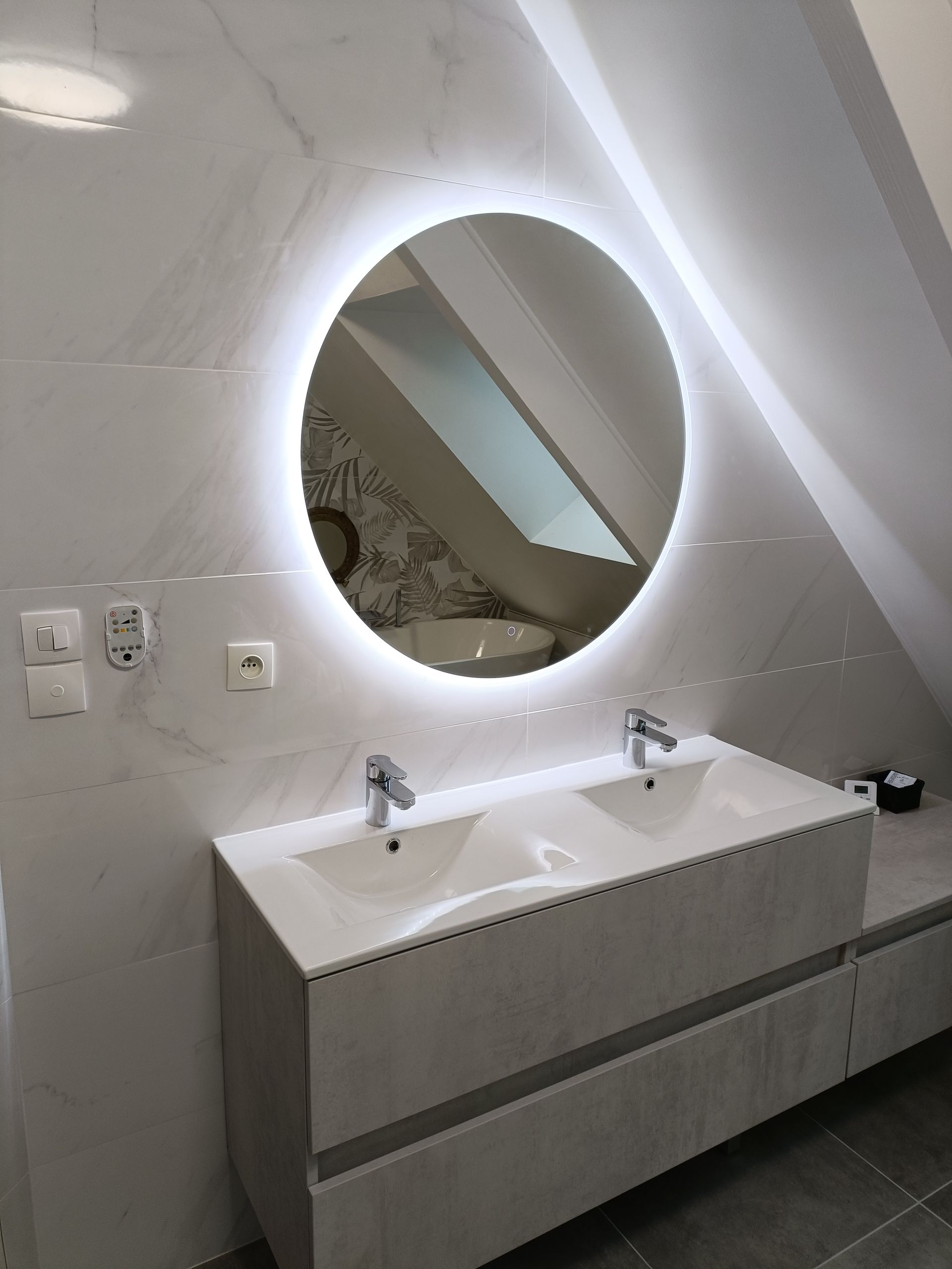 Meuble-lavabo moderne avec double vasque et miroir rond sous un plafond mansardé.