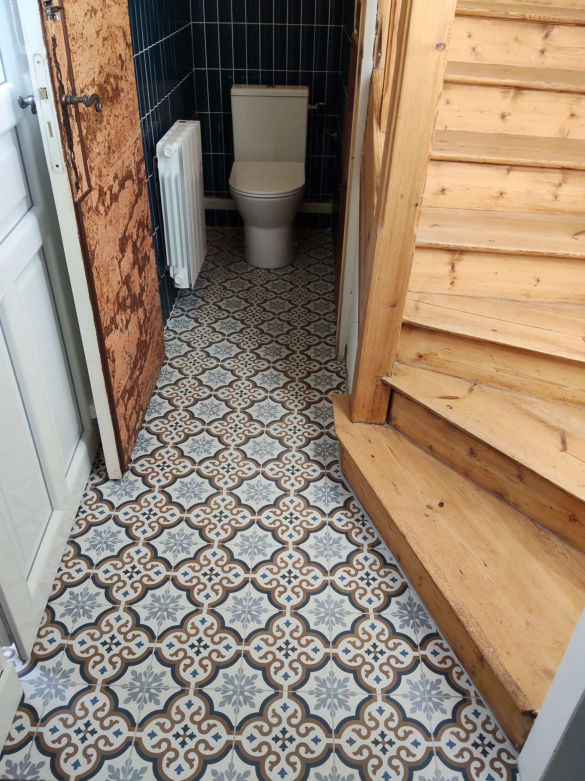 Couloir étroit carrelé menant aux toilettes, avec un escalier en bois à droite et un mur de briques à gauche.