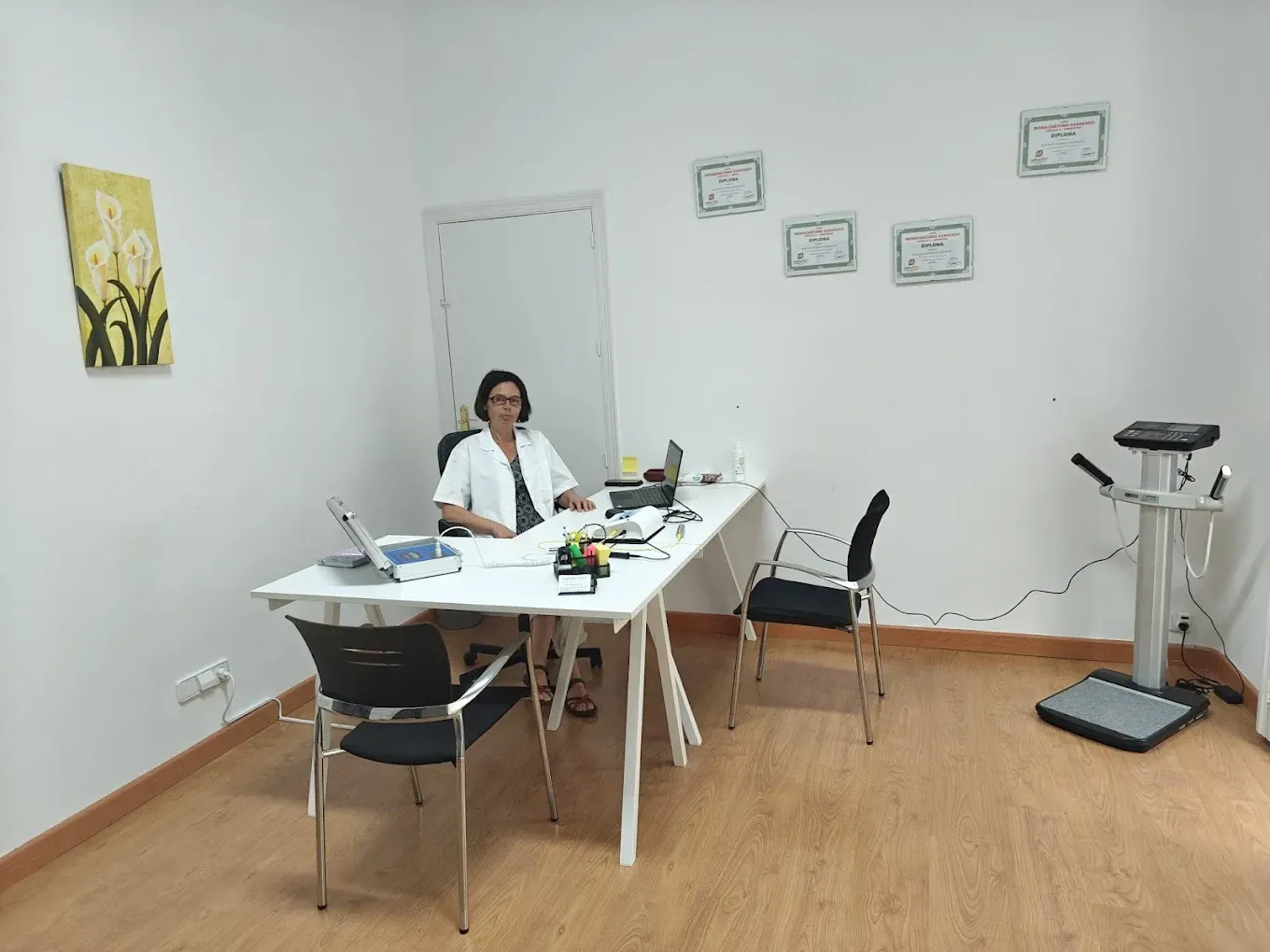 Médico sentado en un escritorio en una oficina con equipo médico. Paredes blancas, suelo de madera y pintura floral.