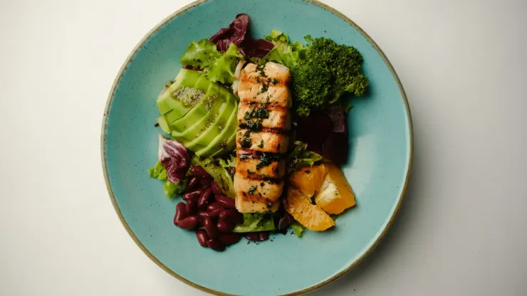 Ensalada con pescado a la parrilla, aguacate, brócoli y naranjas en un plato azul.