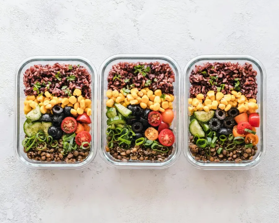 Tres recipientes de vidrio para preparar comidas con capas de granos, garbanzos, verduras y aceitunas.