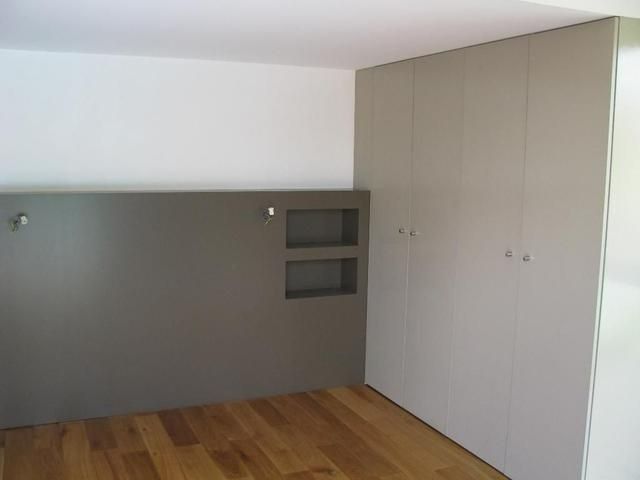 Chambre avec tête de lit grise, étagères intégrées et portes de placard gris clair. Sol en bois.