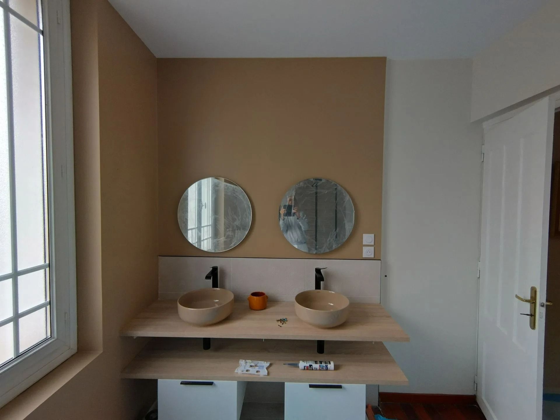 Salle de bains avec deux miroirs ronds au-dessus des lavabos sur un plan de travail en bois. Murs couleur crème, meubles blancs et une fenêtre.