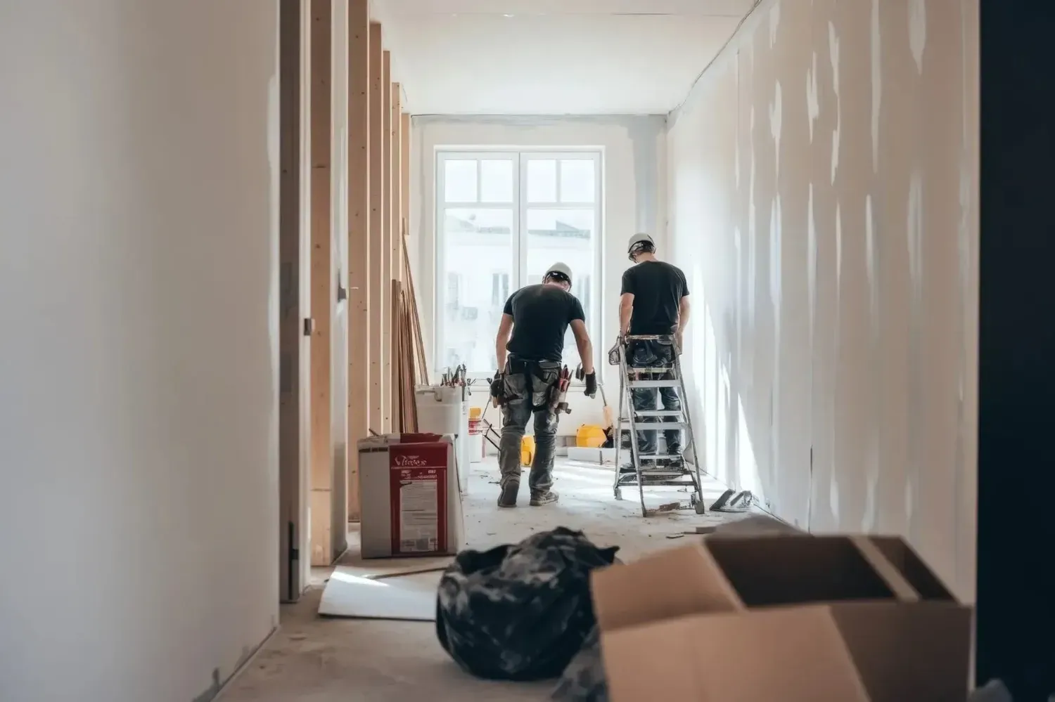 Dos trabajadores en un pasillo luminoso, instalando paneles de yeso con escaleras y herramientas.