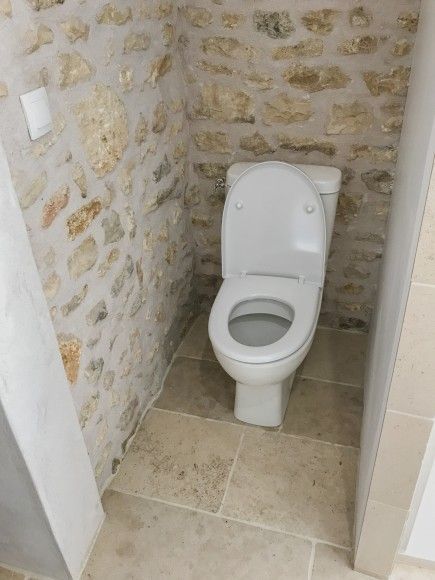 Petites toilettes blanches dans des sanitaires étroits aux murs de pierre et au sol carrelé beige.