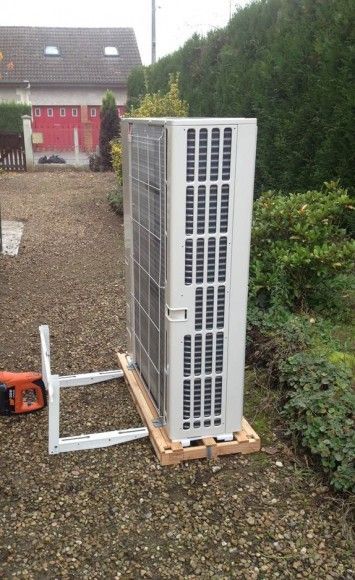 Unité de climatisation extérieure sur une palette en bois dans une cour en gravier, avec un transpalette à côté.