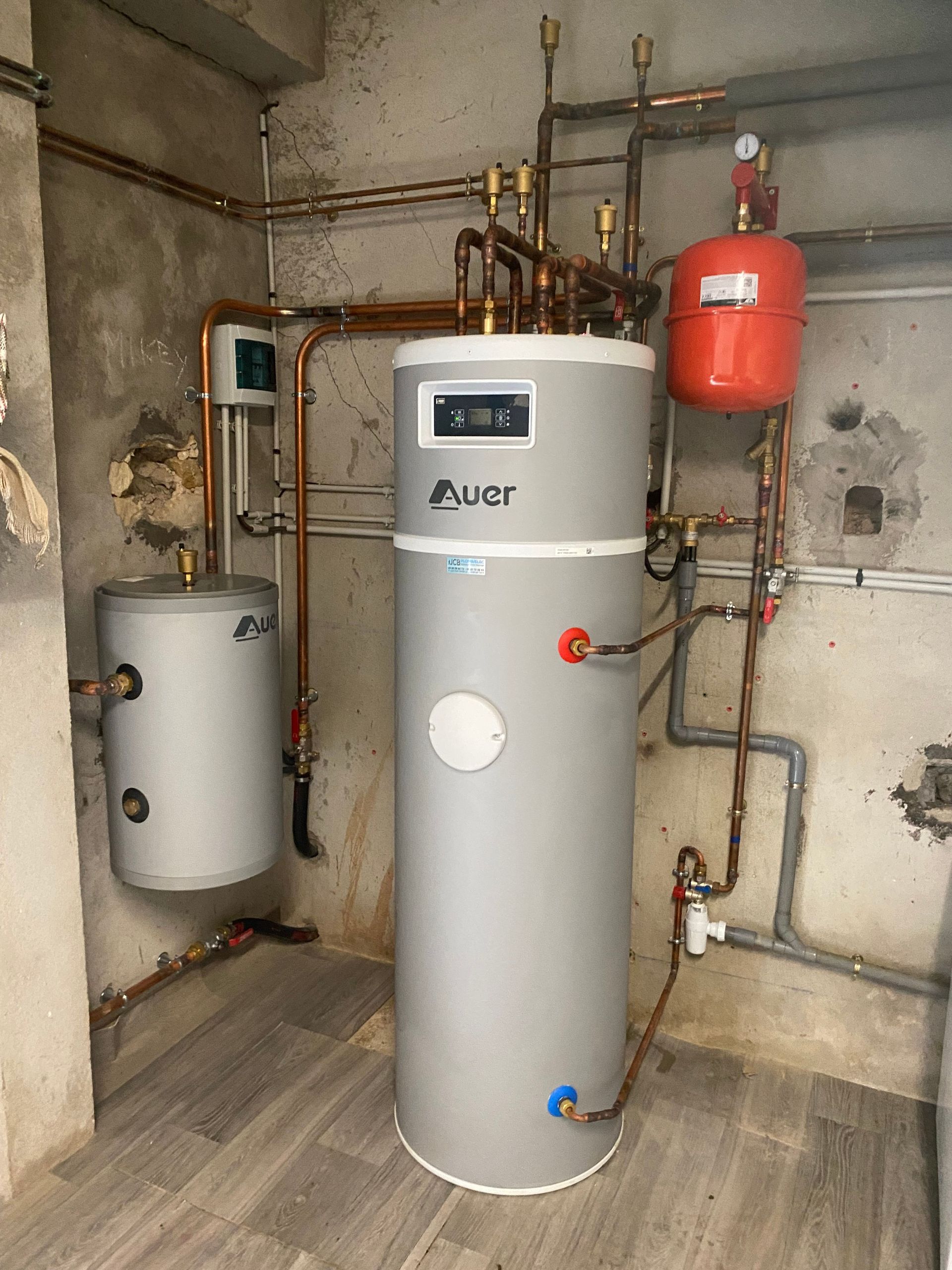 Local technique avec un grand chauffe-eau à eaux grises, un vase d'expansion, un réservoir sous pression rouge et des canalisations apparentes.