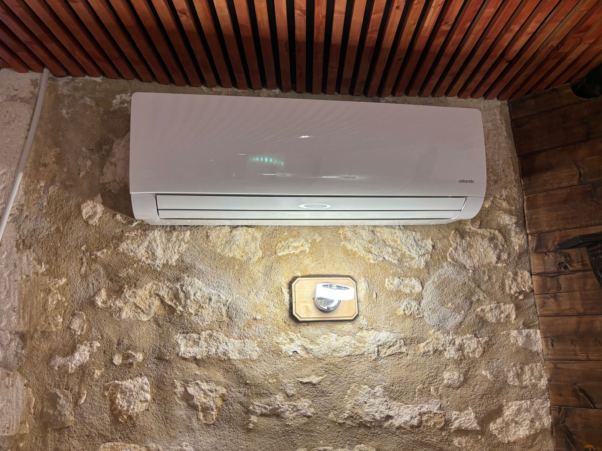 Climatiseur mural blanc au-dessus d'un mur en pierre, avec un petit luminaire en dessous.
