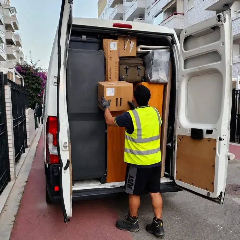 Repartidor con chaleco reflectante cargando cajas de cartón en una furgoneta blanca en la calle.