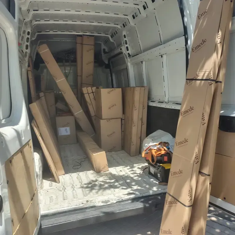 Interior de una furgoneta de carga repleto de cajas de cartón y paquetes largos, con una bolsa de herramientas en el suelo.