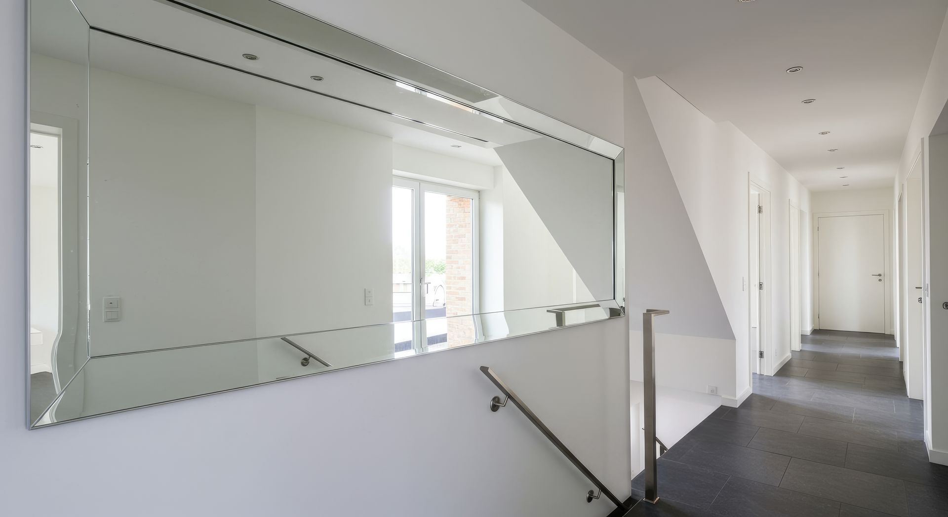 Un couloir moderne avec un grand miroir mural, un escalier à rampe métallique et des murs de couleur claire.