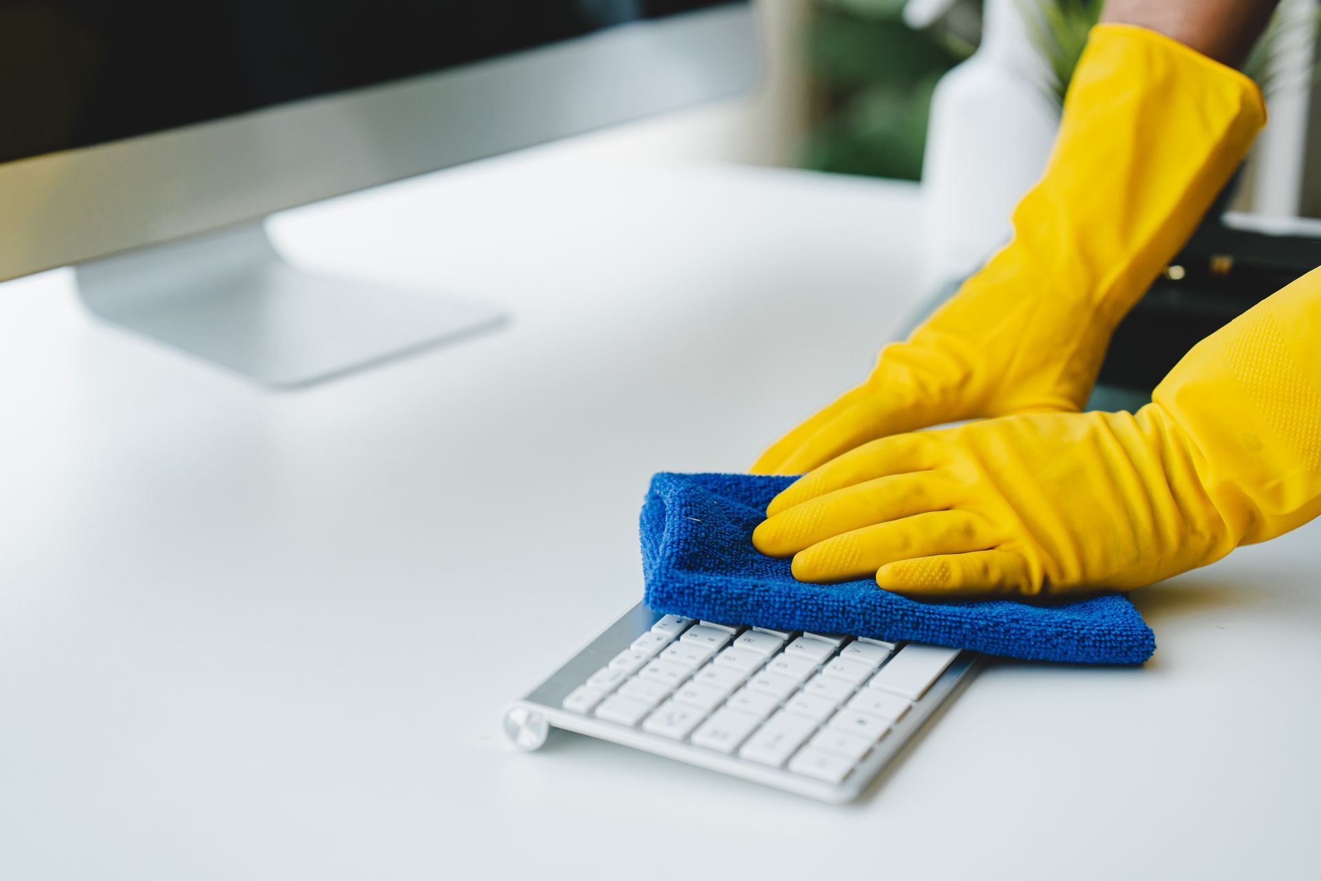 Des mains gantées de gants en caoutchouc jaunes essuient un clavier blanc avec un chiffon bleu sur un bureau blanc.