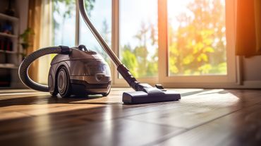 Un aspirateur argenté est posé sur un parquet dans une pièce ensoleillée, avec une grande fenêtre en arrière-plan.