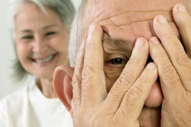 Un anciano se cubre los ojos con las manos mientras una mujer sonríe en el fondo.