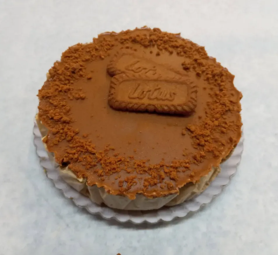 Postre redondo de caramelo coronado con una galleta Lotus, sobre un papel blanco, con galleta desmenuzada alrededor del borde.
