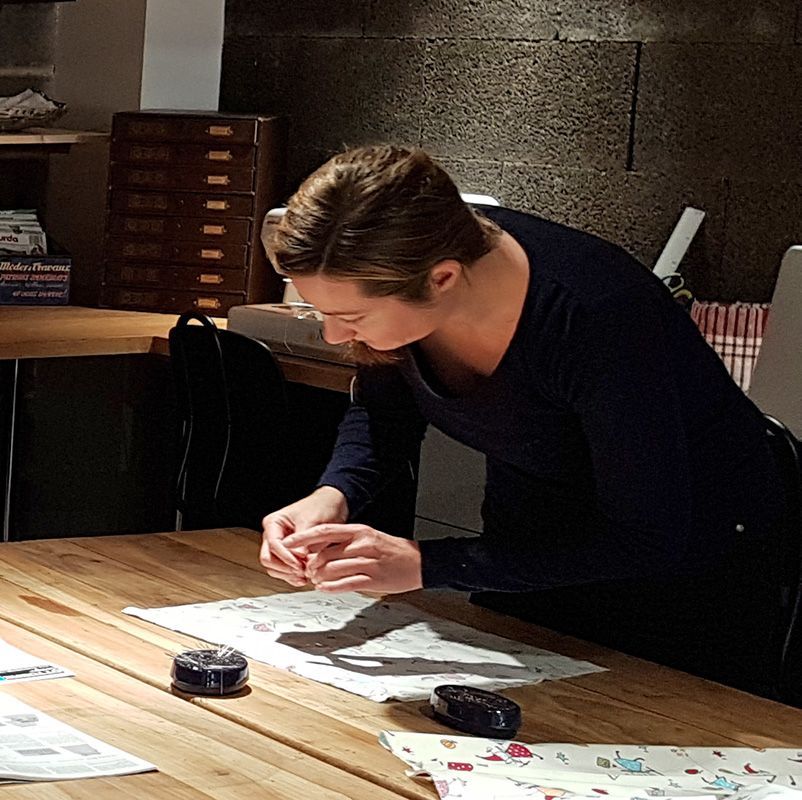 Une femme se penche sur une table en bois, travaillant avec un petit objet sur un tissu à motifs.