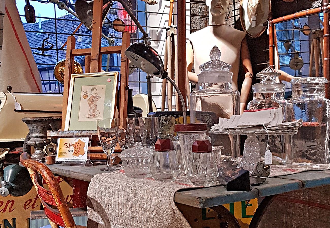 Vitrine d'un magasin d'antiquités : bocaux en verre, tableaux encadrés, mannequin et divers objets vintage disposés sur une table.