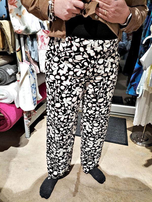 Une personne portant un cardigan marron et un pantalon à fleurs noir et blanc se tient dans un magasin de tissus.