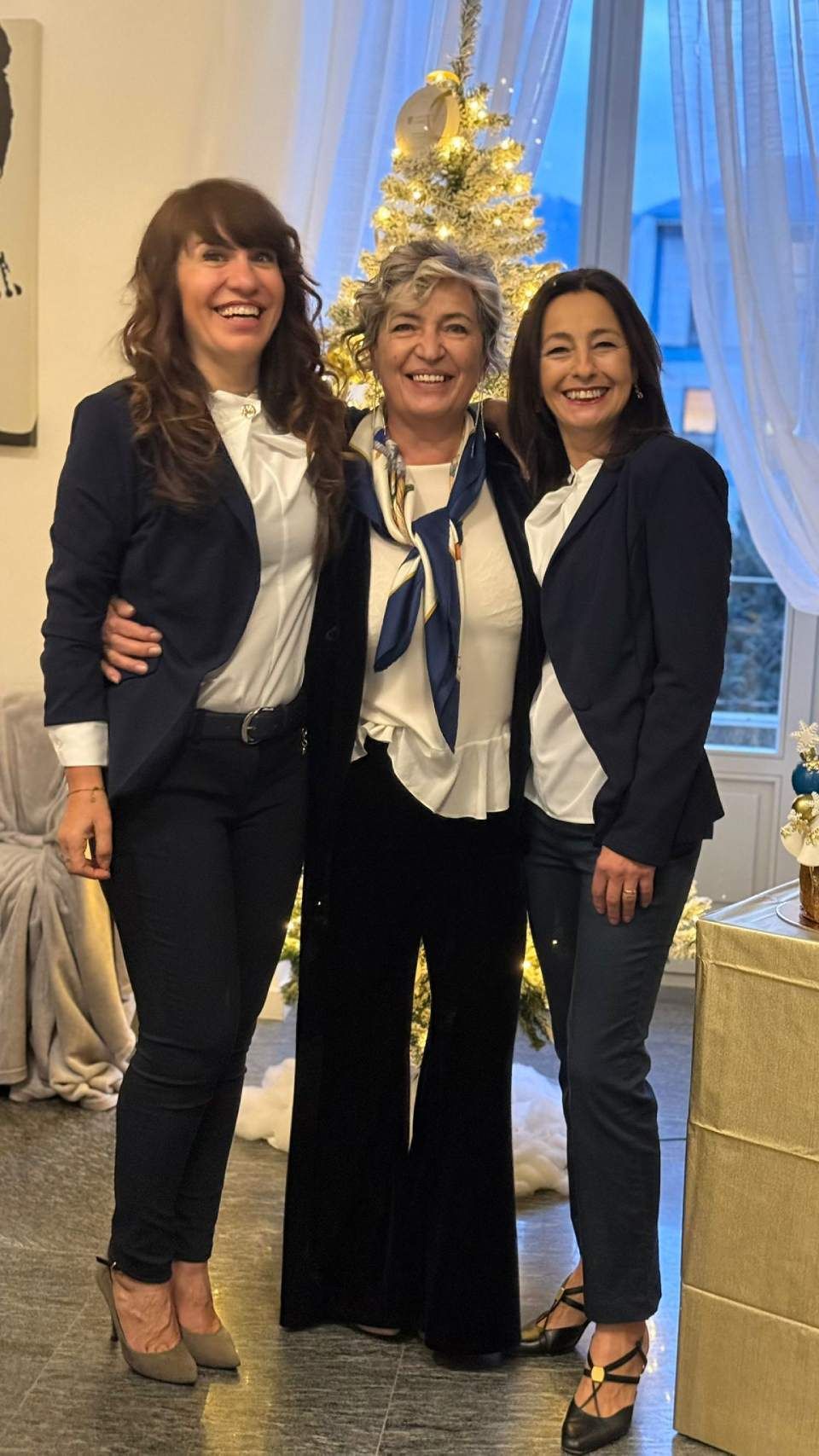 Tre donne sorridenti posano accanto a un albero di Natale decorato, tutte con top bianchi e blazer blu scuro.