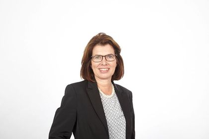 Lächelnde Person in schwarzem Blazer und gemusterter Bluse vor weißem Hintergrund