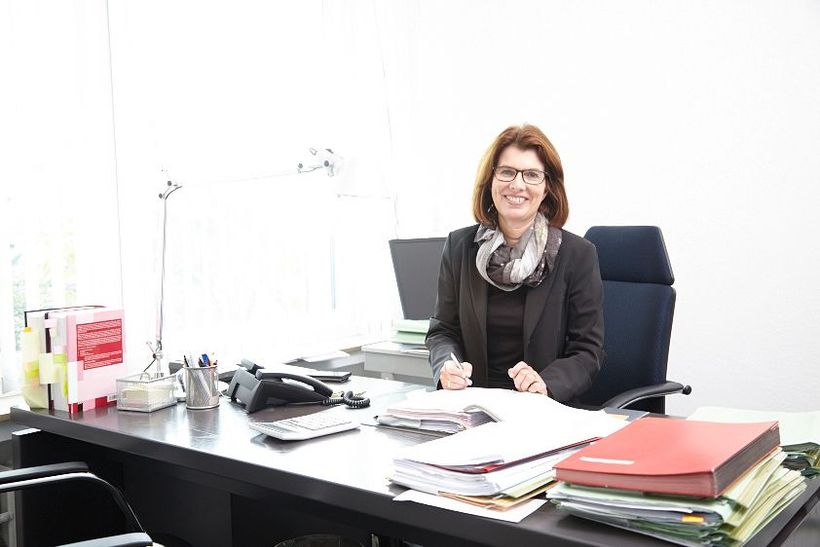 Frau sitzt an einem Schreibtisch mit Papieren, Akten und einem Computer in einem hellen Arbeitsbereich