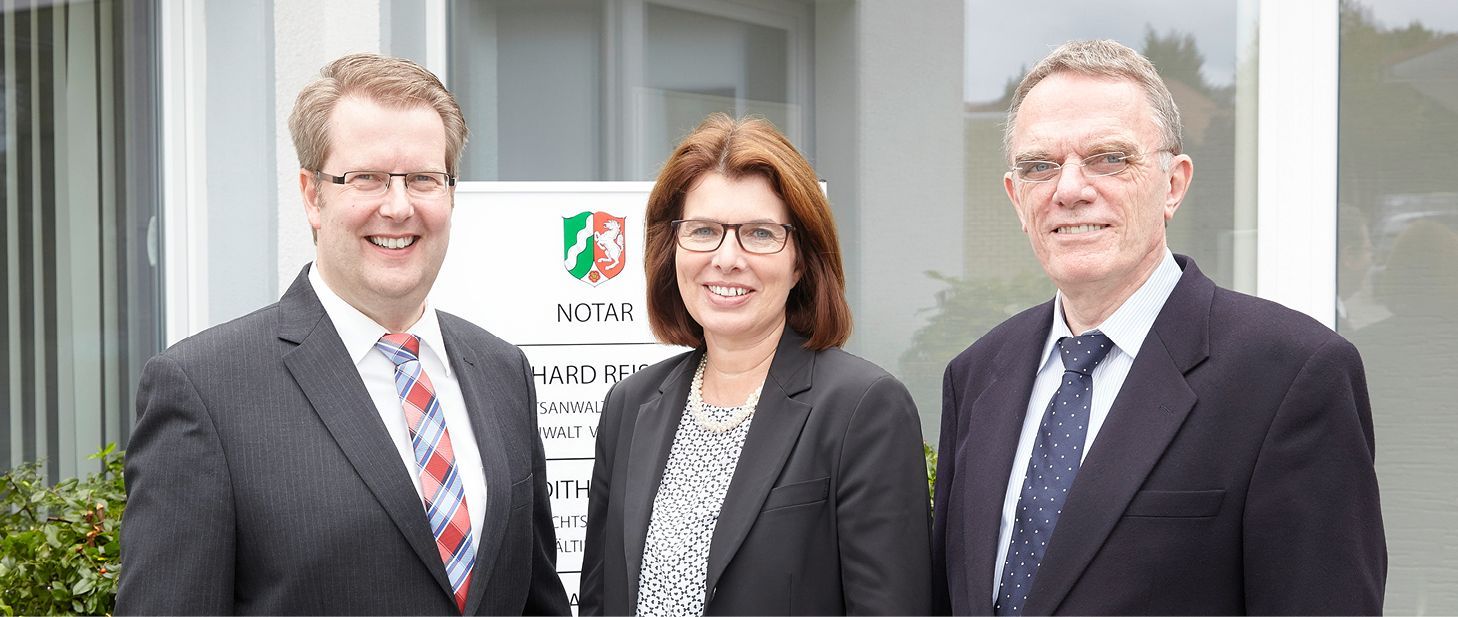 Drei Personen in Geschäftskleidung stehen vor einem Gebäude und lächeln in die Kamera.
