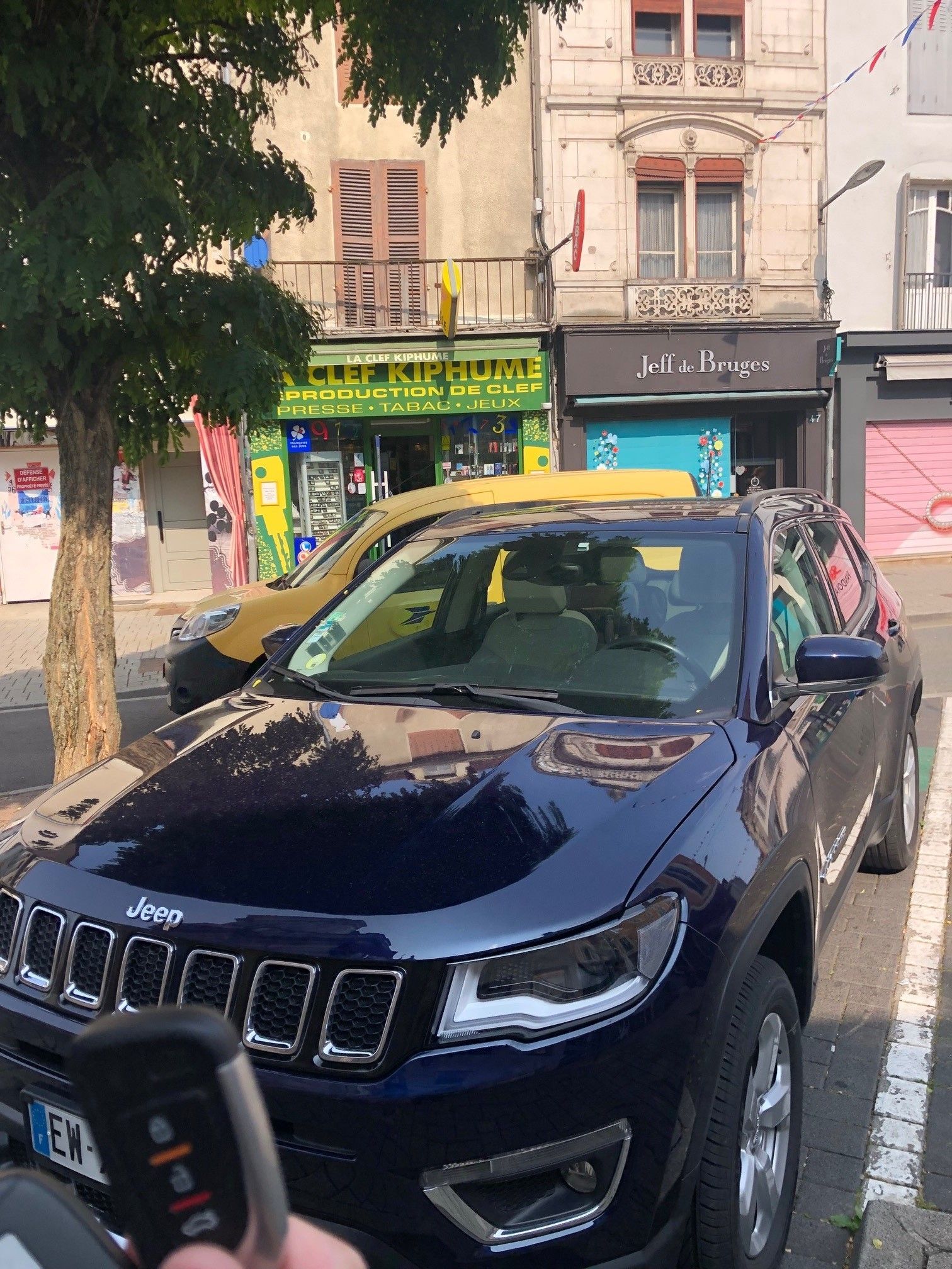 Une Jeep Compass bleue est garée devant des bâtiments, avec sa clé de transport à la main.