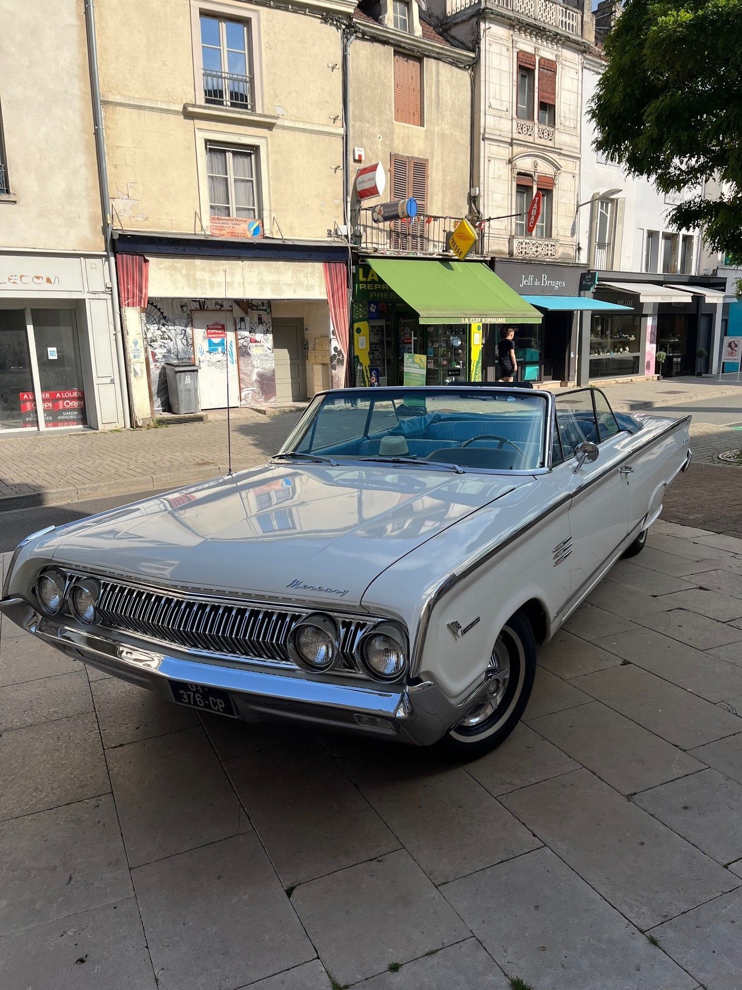 Une décapotable blanche vintage garée sur une place pavée devant des magasins.