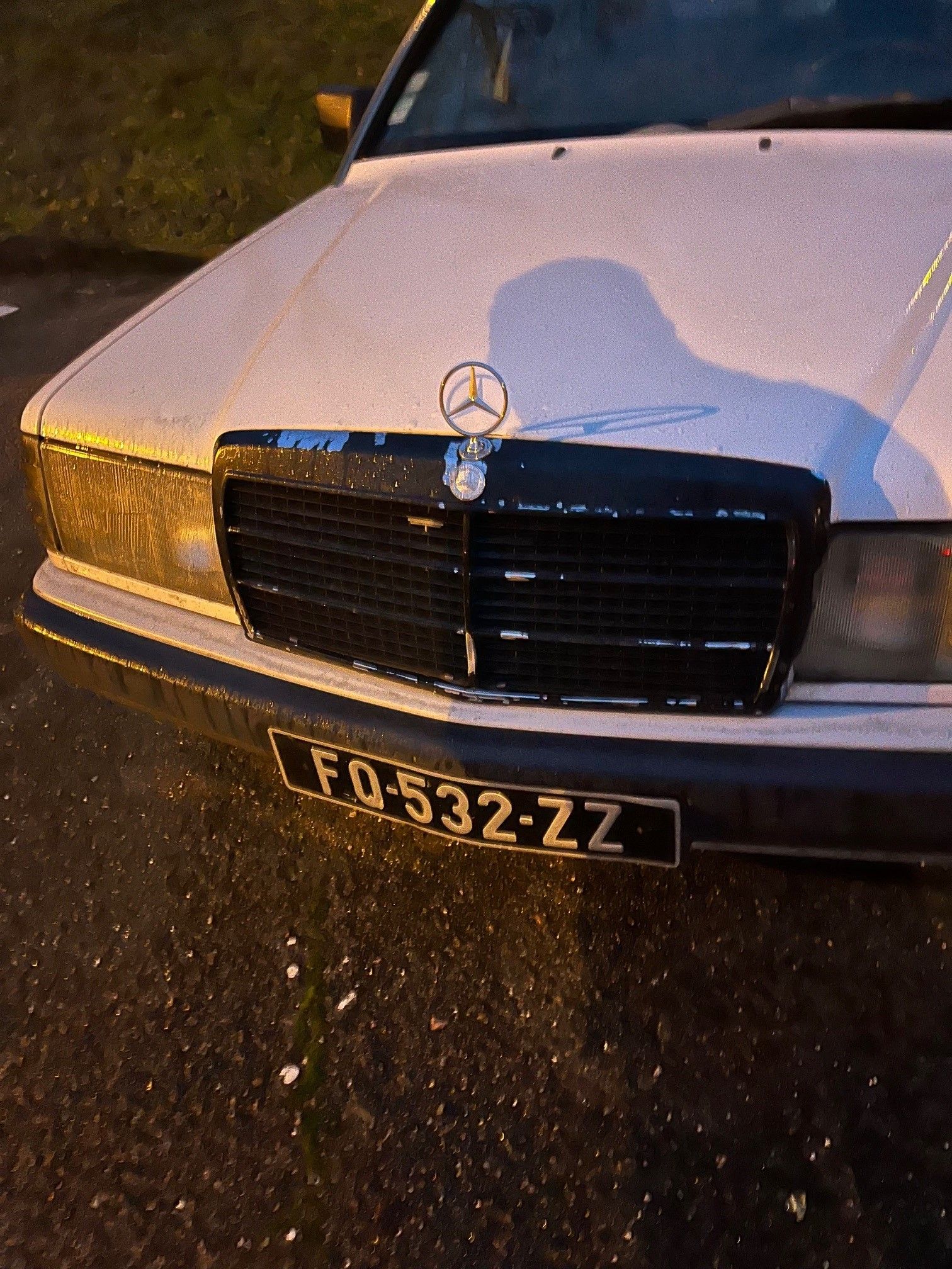 Berline Mercedes-Benz blanche avec calandre noire et plaque d'immatriculation « FQ-532-ZZ » dans un environnement humide.
