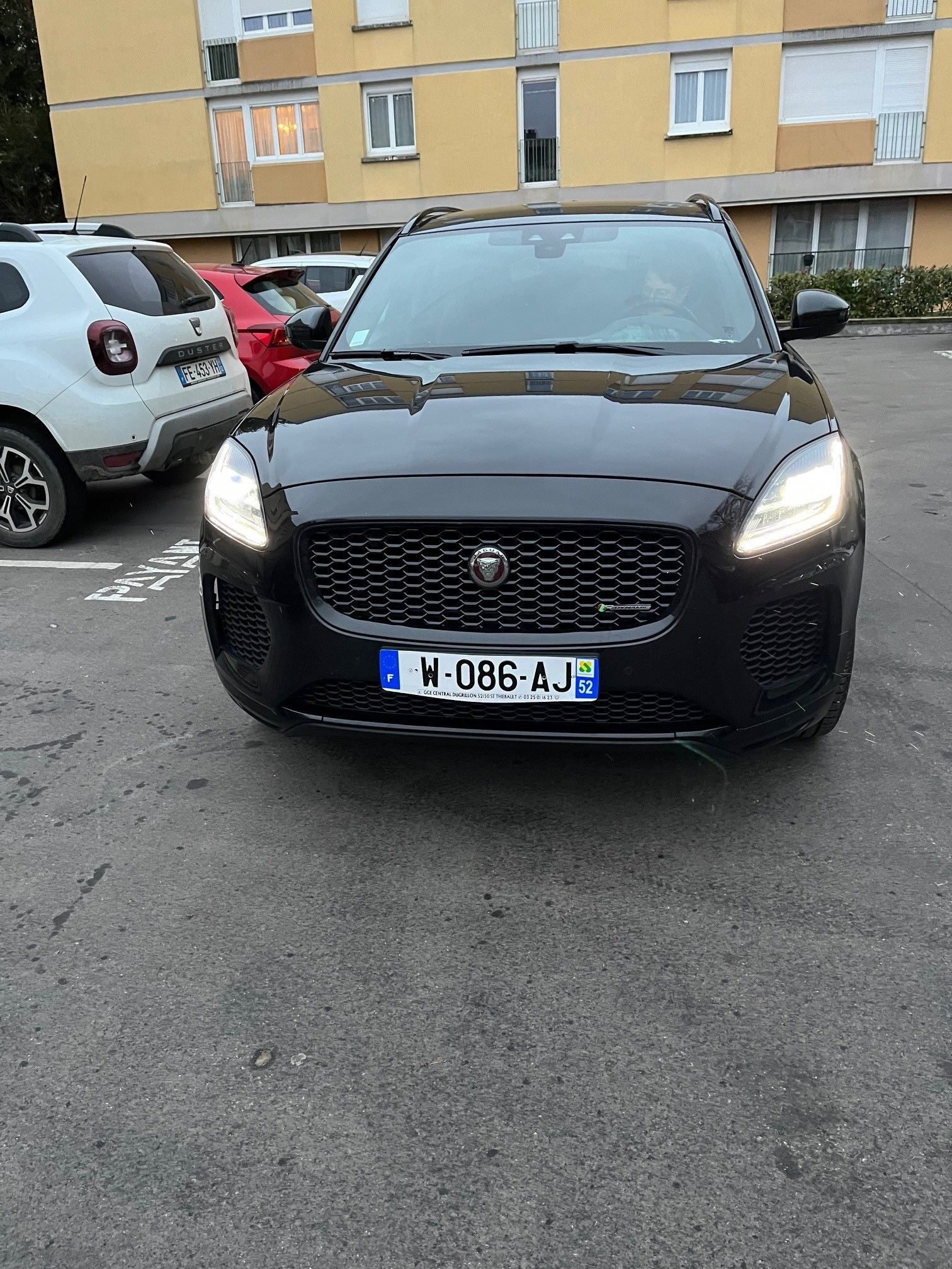 Jaguar E-Pace noire garée devant un bâtiment. Phares allumés, plaque d'immatriculation visible.