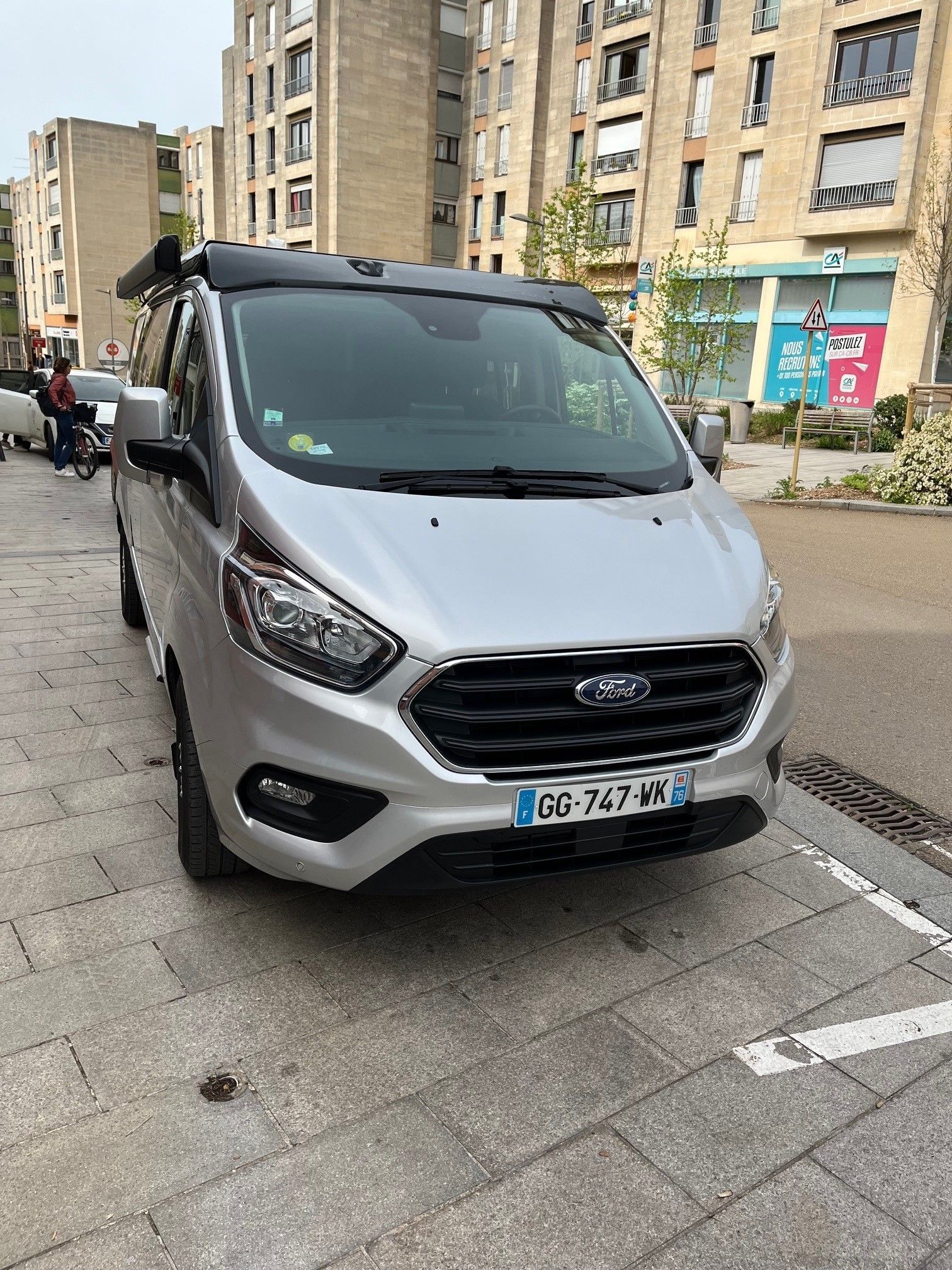 Fourgonnette Ford argentée garée dans une rue pavée de briques, avec des bâtiments en arrière-plan. Plaque d'immatriculation française.