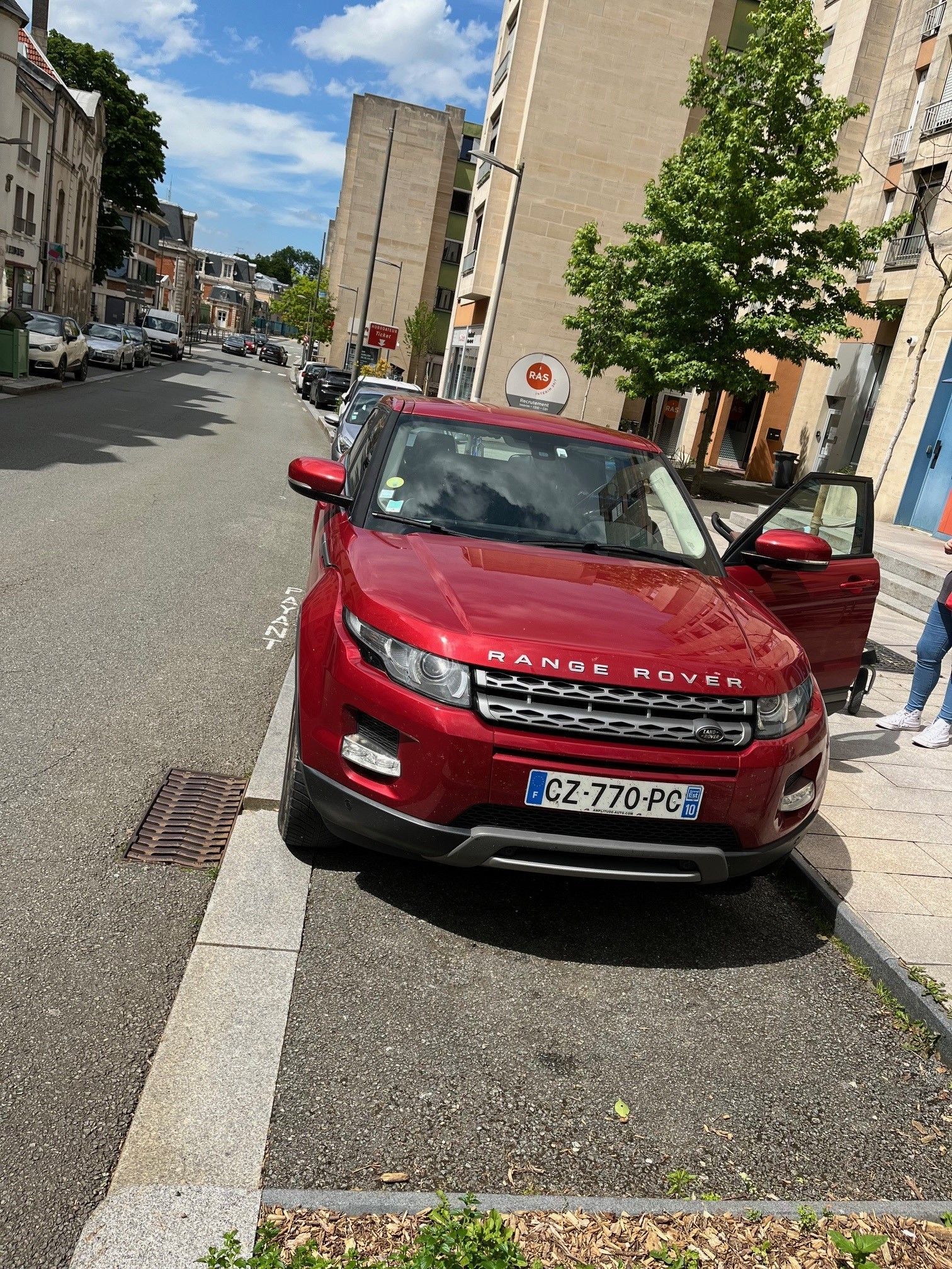 Un Range Rover rouge est stationné dans une rue de la ville ; la portière passager est ouverte. Des bâtiments et des voitures stationnées bordent la route.