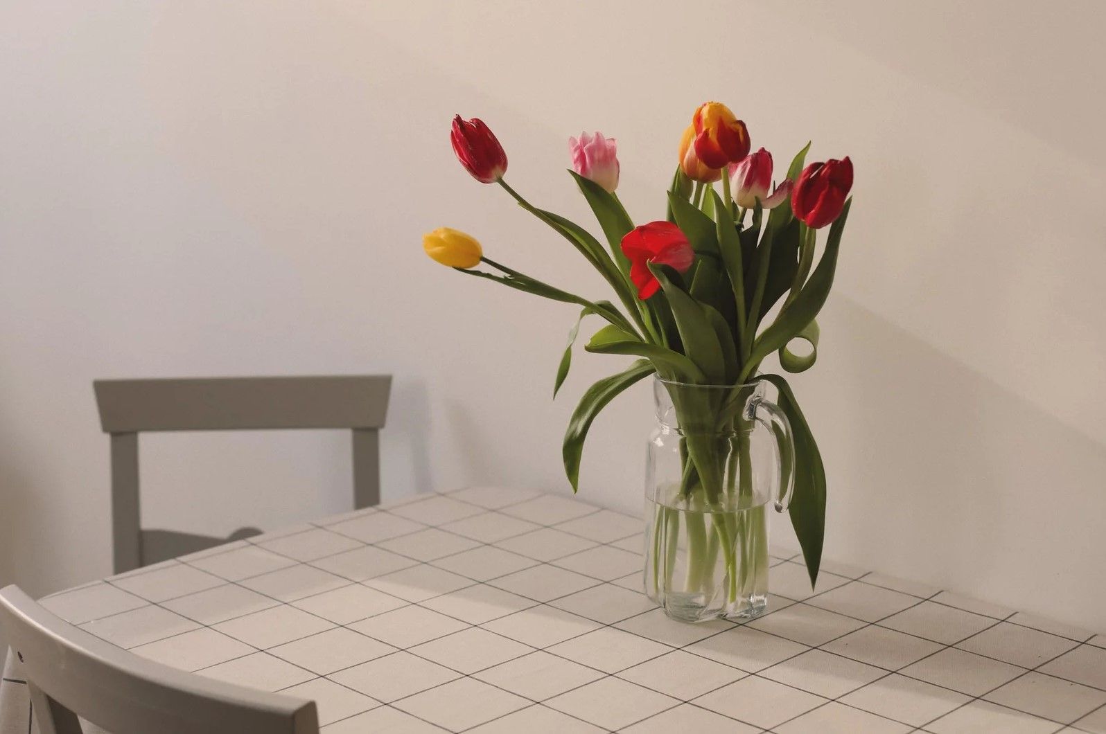 Un jarrón de tulipanes de colores reposa sobre una mesa frente a una silla gris; pared blanca.