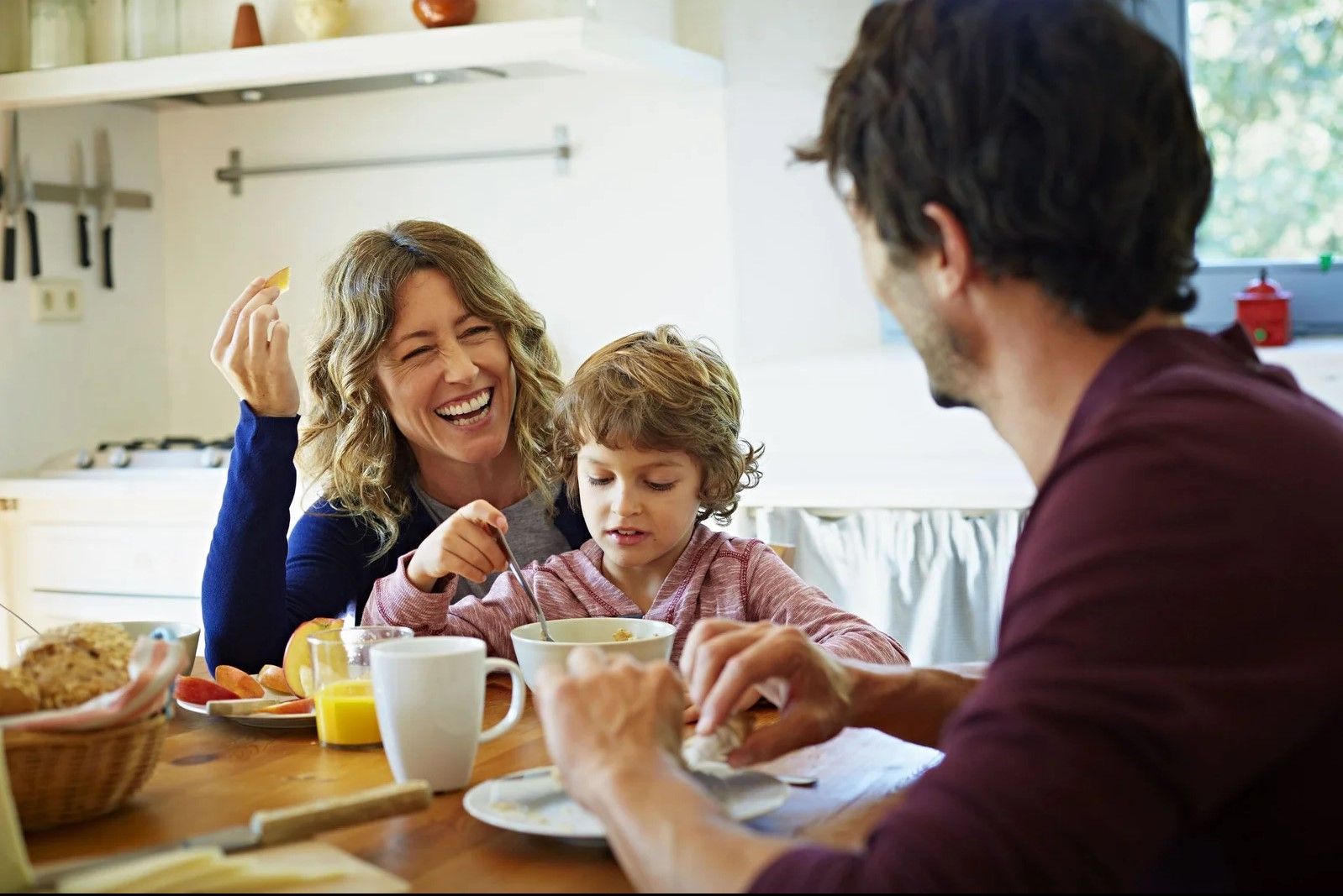 Una familia ríe y desayuna junta en una mesa de la cocina. La madre sonríe y el niño come cereal.