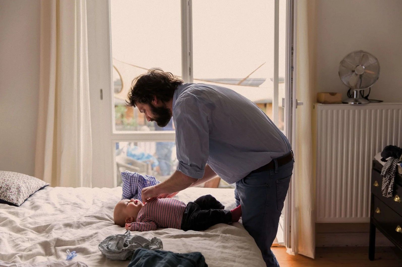 Un hombre cambia la ropa de un bebé en una cama de un dormitorio luminoso. Se ve una ventana con un ventilador y un radiador en la pared.
