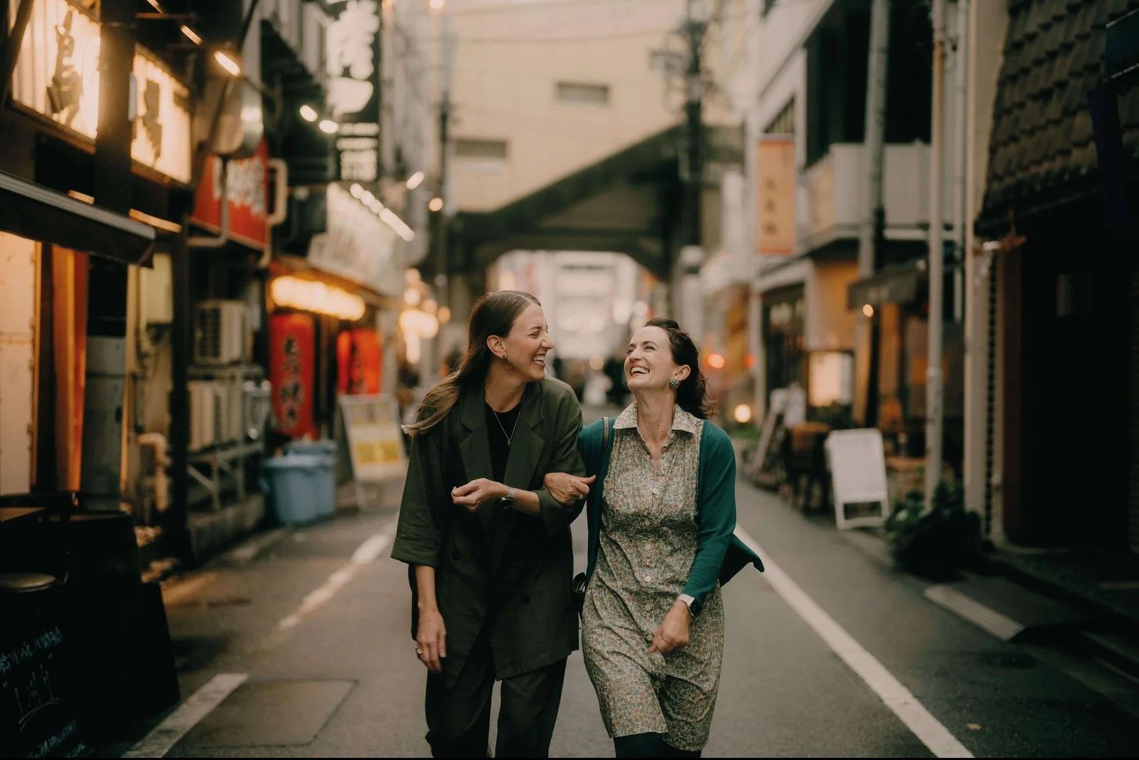 Dos mujeres del brazo, sonriendo y caminando por una calle estrecha llena de tiendas, probablemente en una ciudad.