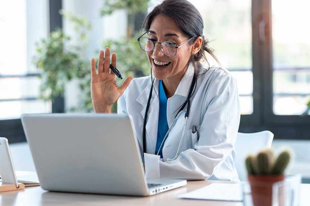 Un médico sonriente con una bata blanca saluda durante una videollamada en una computadora portátil, en un interior con plantas y un cactus.