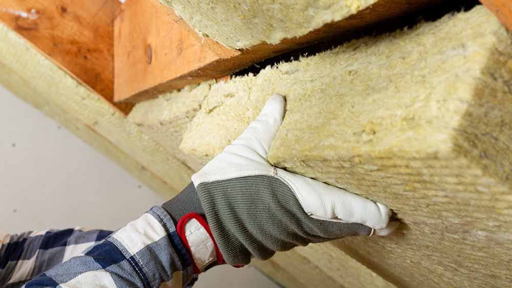 Behandschuhte Hand drückt weißen Dämmschaum unter Holzbalken.