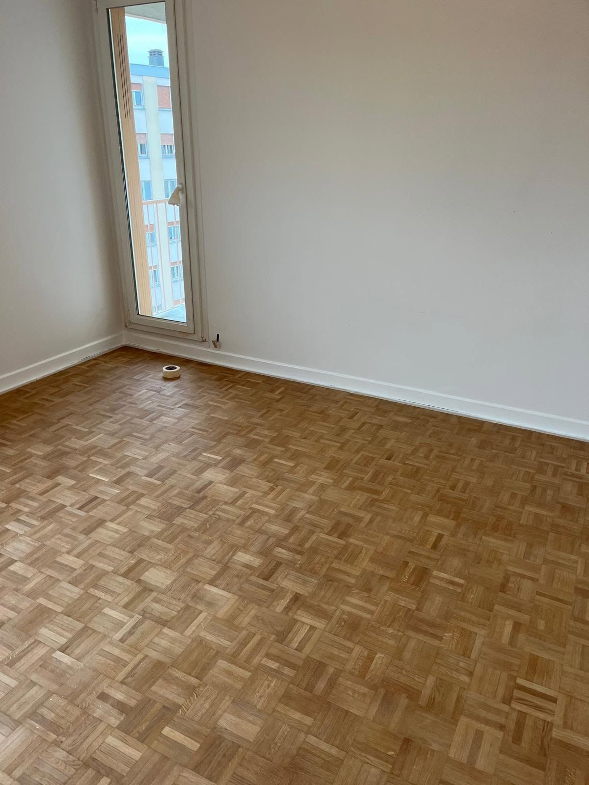 Pièce vide avec parquet, murs blancs et une fenêtre étroite laissant entrer la lumière du jour.