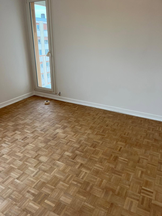 Pièce vide avec parquet, murs blancs et une fenêtre étroite laissant entrer la lumière du jour.