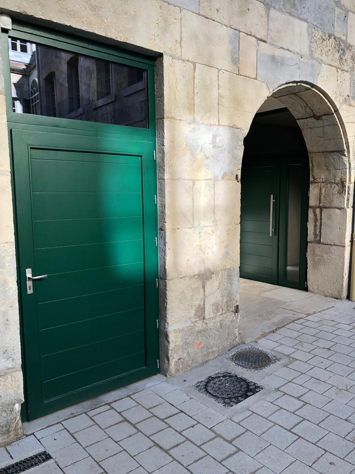 Porte verte et entrée voûtée en pierre donnant sur un trottoir pavé.