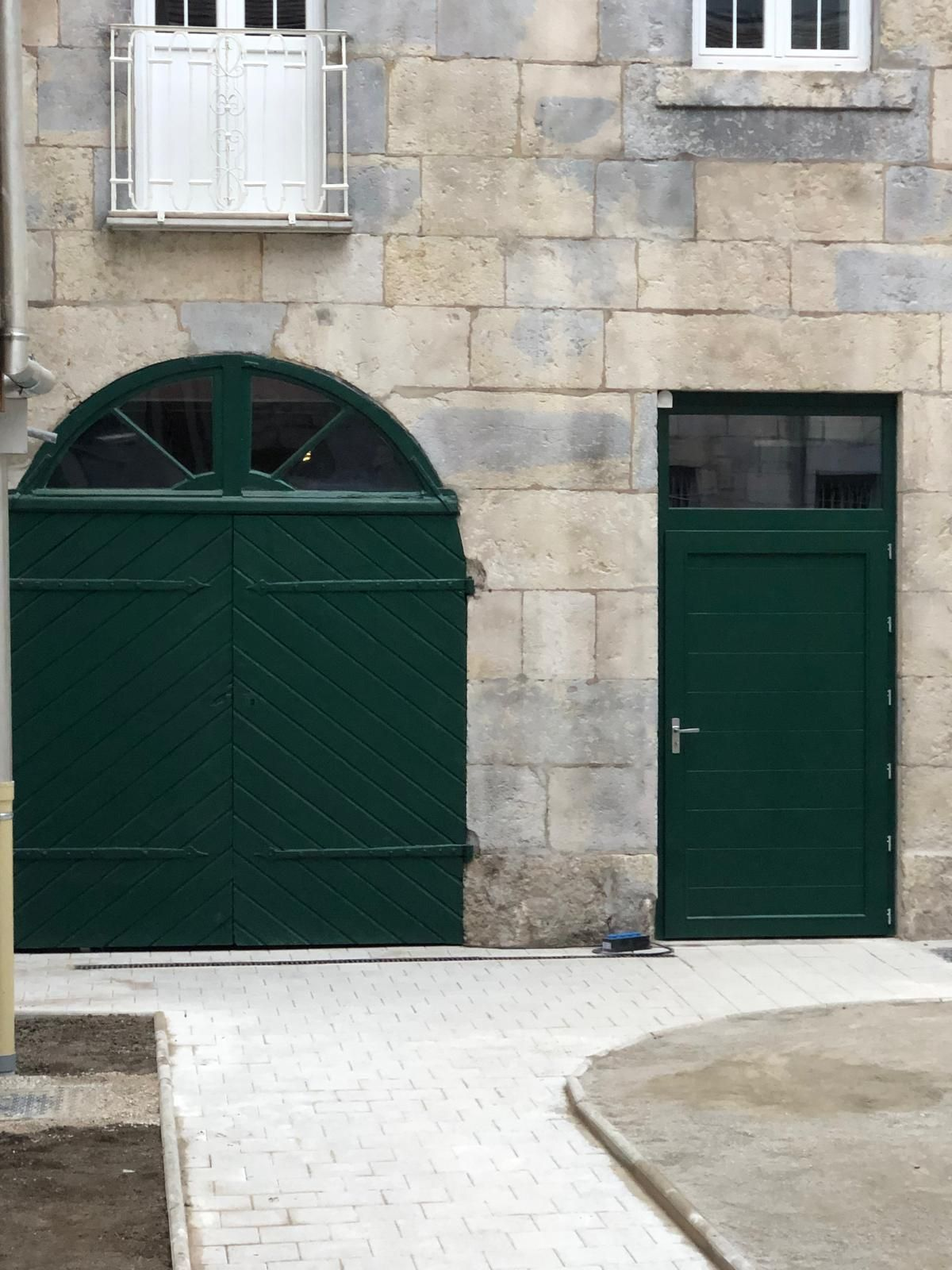 Porte de garage cintrée verte et entrée latérale intégrées à une façade de bâtiment en pierre.