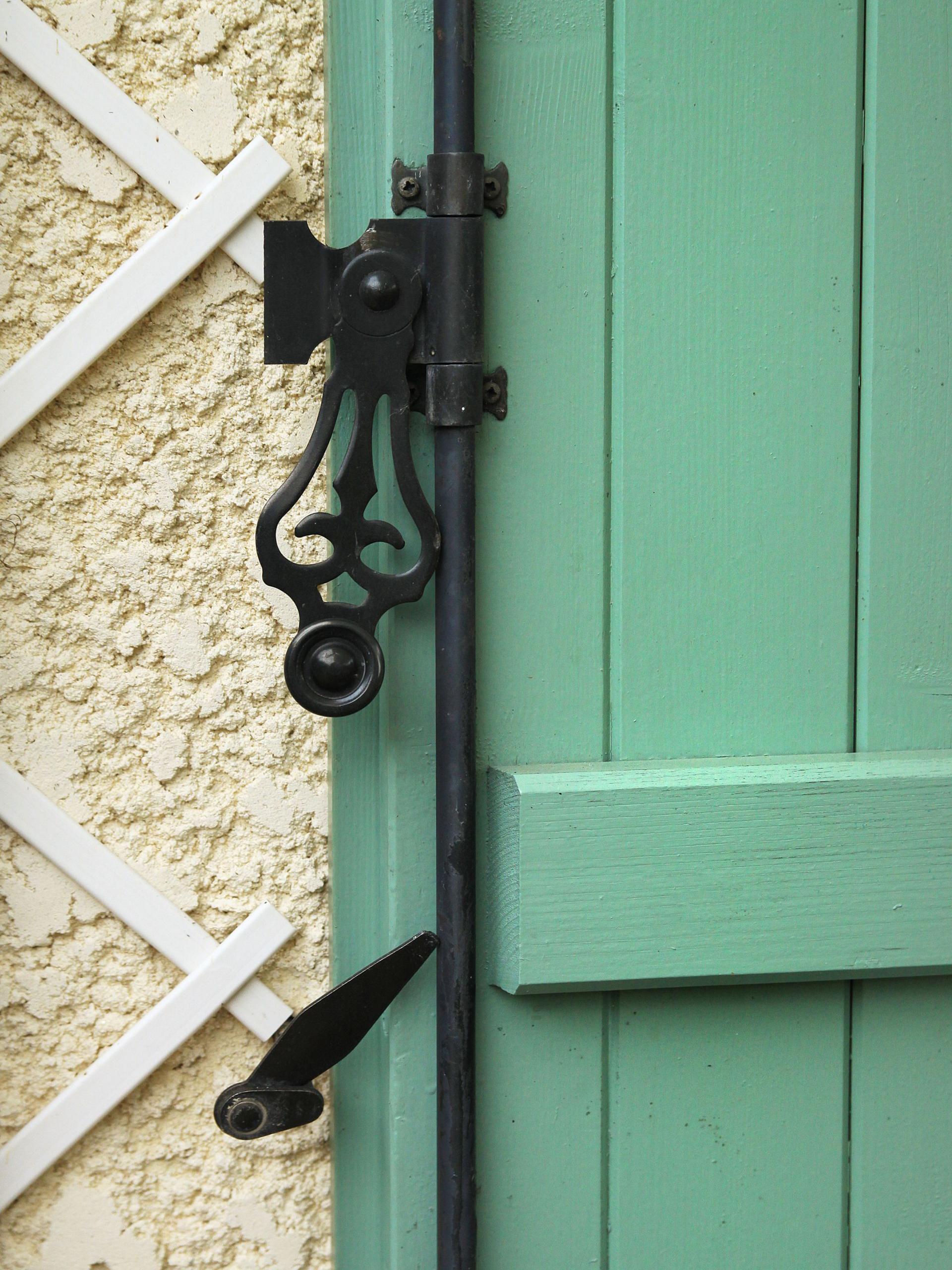 Loquet en fer noir sur un volet en bois vert menthe, à côté d'un mur en crépi blanc.