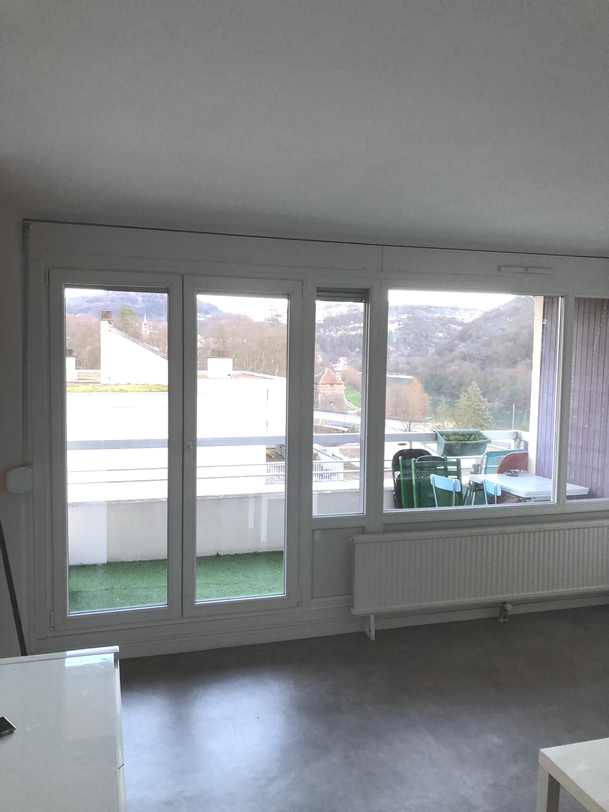 Salon vide avec portes vitrées, radiateur et vue sur un balcon et les collines environnantes.